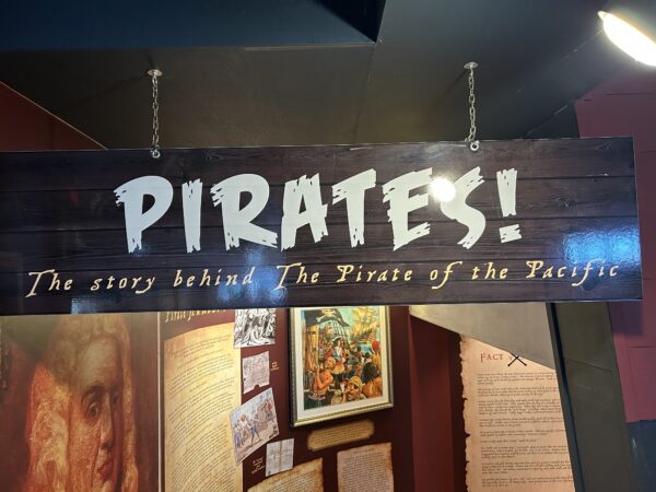 A faux wood sign hanging from two chains that reads "Pirates! The Story Behind the Pirate of the Pacific"