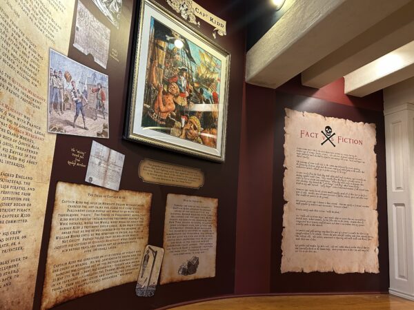 An exhibit wall painted dark red displaying classic artwork of pirates and several text panels describing the history of piracy. The panels are designed to look like worn parchment evoking a treasure map.