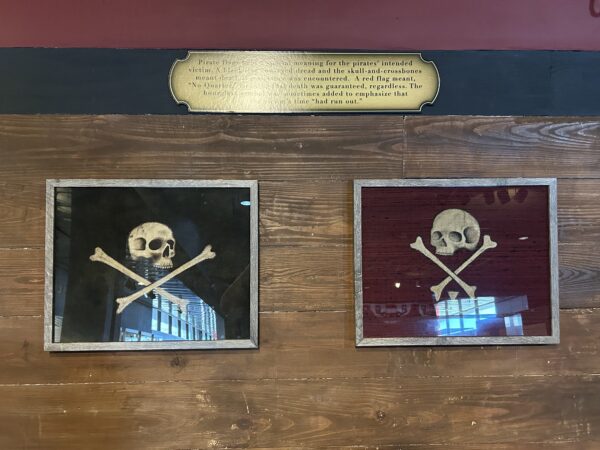 A pair of Jolly Roger flags. Both are decorated with the skull and crossbones motiff, although the flag on the left is made of black fabric and the skull is looking to the right. The flag on the right is made of red fabric with the skull looking to the left and a white hourglass shape included beneath the crossed bones.